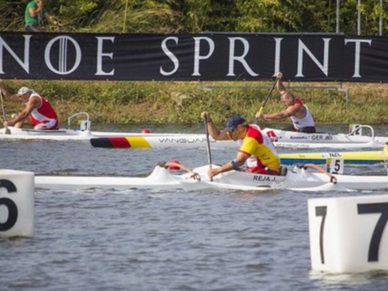 Competencia de canoas sprint en un río, con dos canoístas en el agua y un banner que dice 'CANOE SPRINT &'.