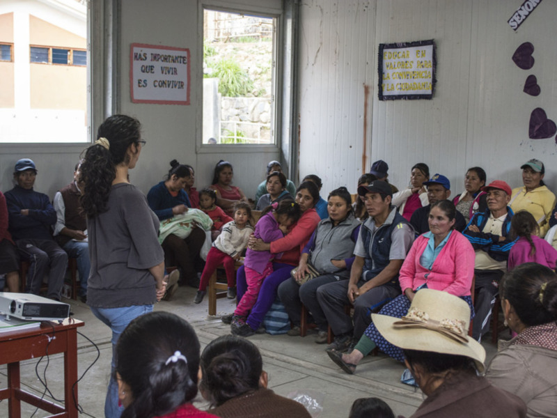 Reunión comunitaria en un espacio educativo, con una mujer dando una charla a un grupo de personas sentadas. Carteles en español promueven valores y convivencia.