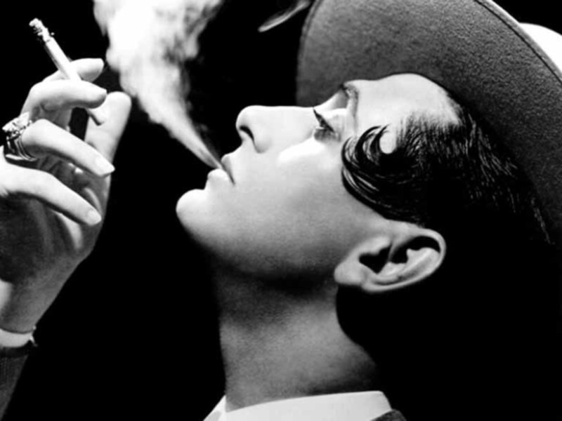 Retrato en blanco y negro de una persona fumando, con sombrero y expresión serena.