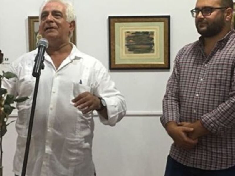 Hombre hablando en un micrófono en una exposición de arte, junto a otro hombre en una sala con cuadros enmarcados.
