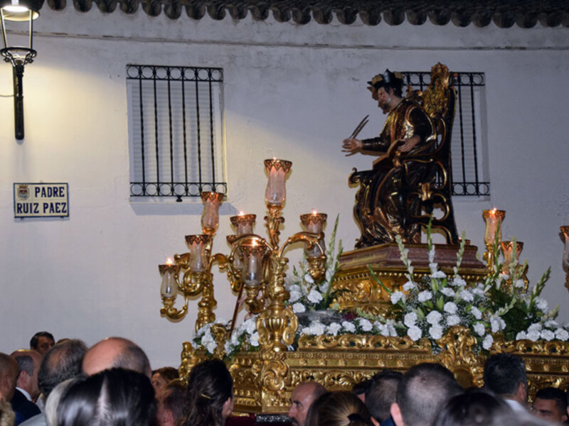 Estatua dorada en un altar con flores y velas, junto a un letrero 'Padre Ruiz Paez' en la pared. La escena se desarrolla en un entorno religioso, probablemente durante una procesión o ceremonia.