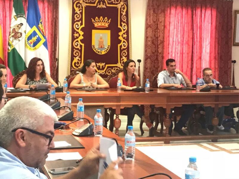 Sala de reuniones con personas sentadas en una mesa, con banderas y un escudo en el fondo.