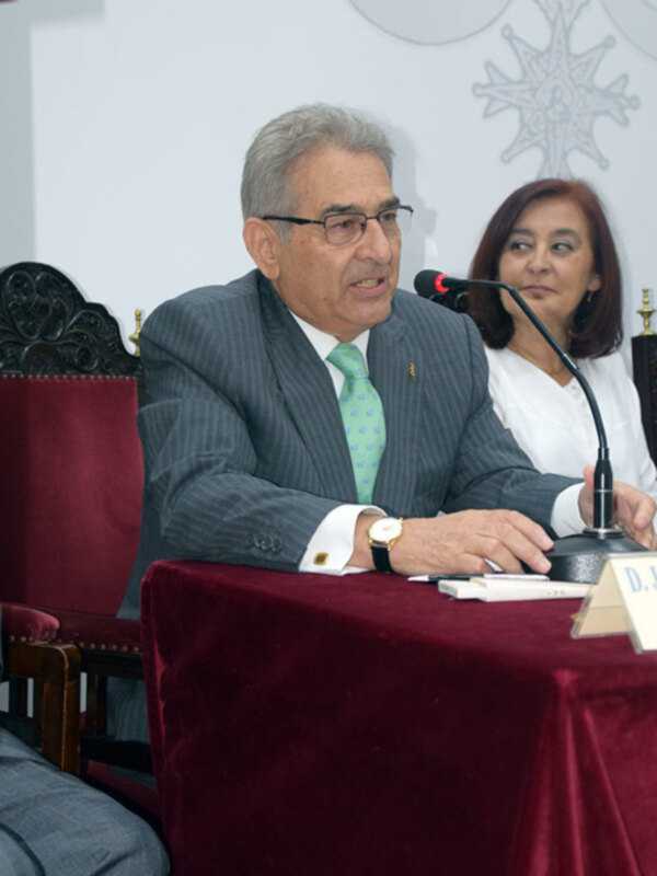 Un hombre en traje y corbata verde se dirige a un micrófono en un evento formal, con una mujer detrás de él. La escena se centra en la comunicación y la presentación en un ambiente formal.