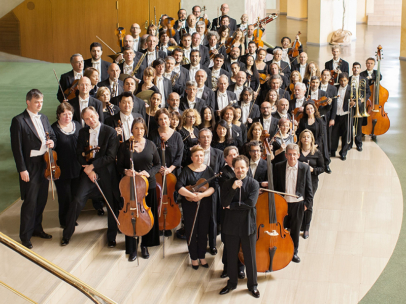 Orquesta de música clásica en un escenario bien iluminado, con instrumentistas en trajes formales y instrumentos de cuerda y viento.