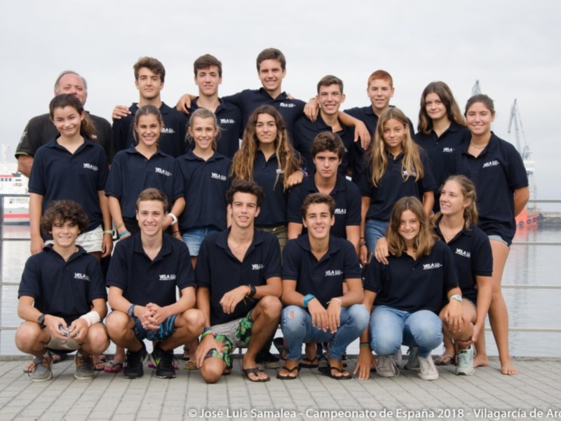 Equipo de jóvenes en camisetas azules posando en la orilla del mar, con un fondo de agua y un barco en el horizonte. La imagen es de la Copa de España de Vela 2018 en Vila de Arousa, Galicia, España.