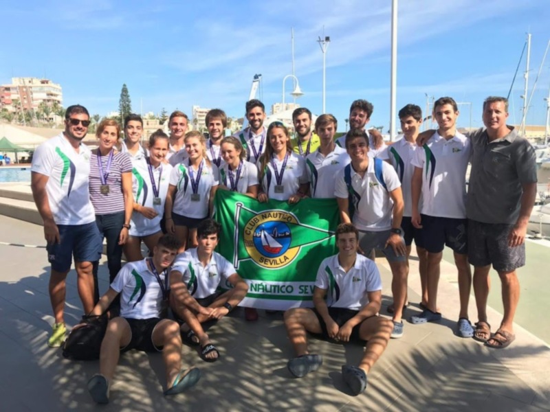 Equipo de natación celebrando un logro deportivo en el puerto deportivo de Sevilla, con medallas y una bandera del Club Náutico Sevilla.