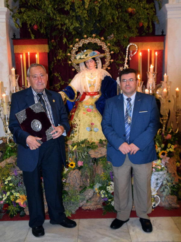 Dos hombres en trajes formales posan frente a una imagen religiosa adornada con flores y velas, en un ambiente ceremonial.