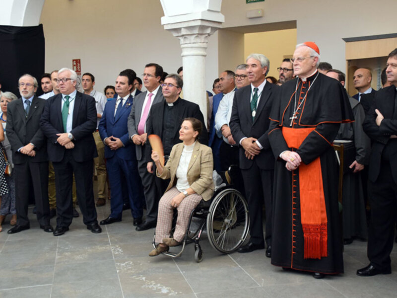 Un grupo de personas, incluyendo un cardenal y otras figuras religiosas y civiles, participa en una reunión o evento formal en un espacio interior bien iluminado.