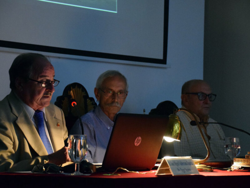 Tres hombres sentados en una mesa con un ordenador portátil HP, en un entorno de conferencia o reunión formal.