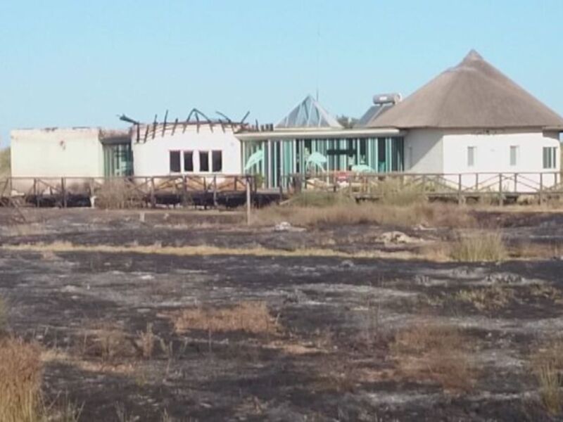 Un edificio de estilo rústico con techo de paja se encuentra en medio de un paisaje desolado, con pasto quemado y un cielo azul claro en el fondo.