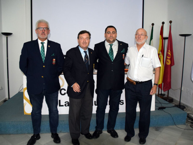Cuatro hombres en trajes posan juntos en un evento formal, con banderas y un cartel en el fondo.