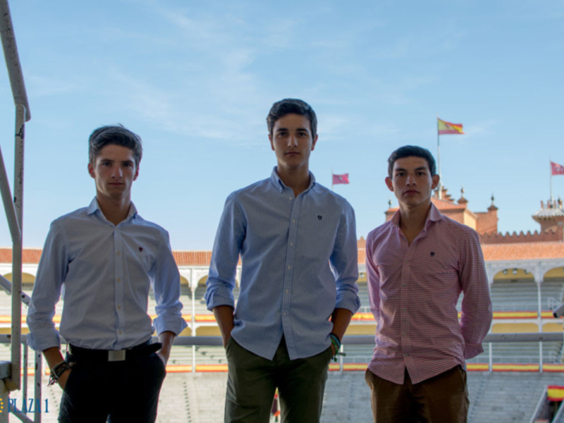 Tres jóvenes en camisas formales frente a una plaza de toros con banderas españolas al fondo.