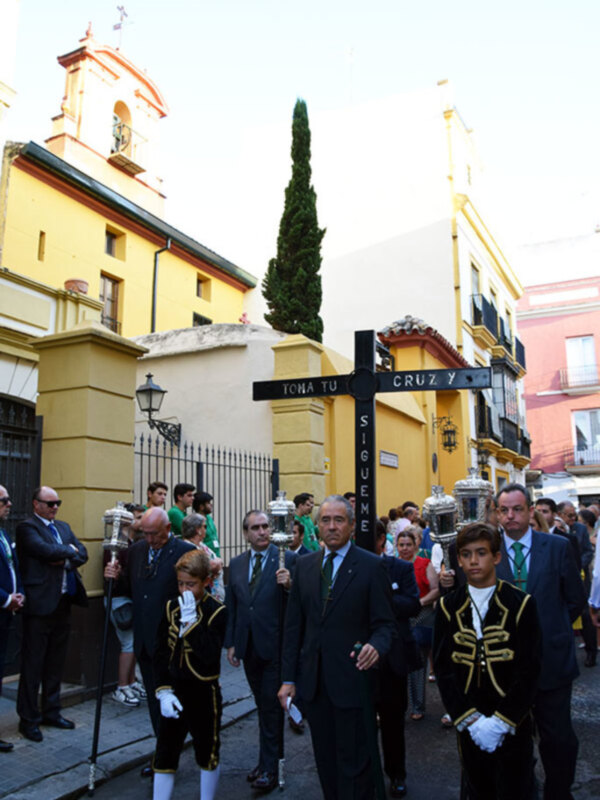 Procesión con personas en trajes formales y niños en uniforme, portando cruces y ornamentos religiosos en una calle de edificios históricos.