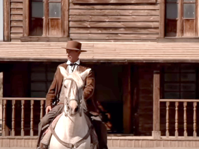 Hombre en traje de vaquero montando a caballo frente a una casa de madera del Oeste.