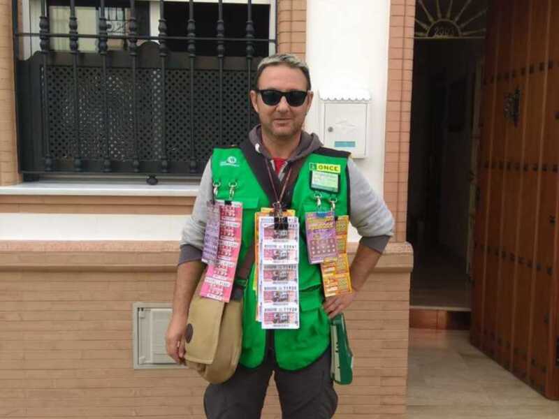 Voluntario de la ONCE con lotería en el pecho, frente a una casa.