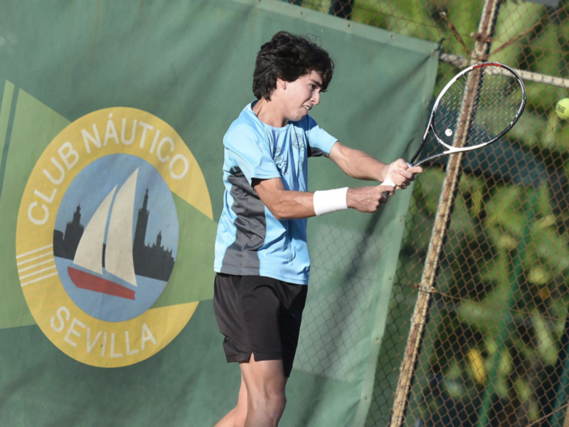 Un joven juega tenis en el Club Náutico Sevilla, con una raqueta y una pelota en el aire. En el fondo, se puede ver una valla verde con el logotipo del club.