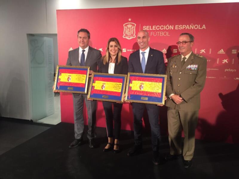 Tres personas posan con sus reconocimientos en una ceremonia de la Selección Española de Fútbol, frente a un fondo rojo con la bandera española y el escudo de la selección.