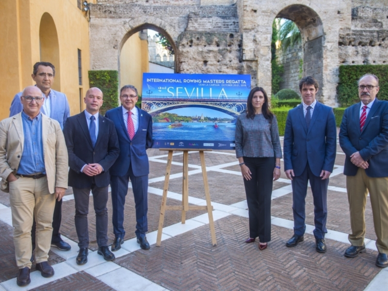 Presentación de la Regata de Remo Masters en Sevilla, con un grupo de personas frente a un cartel que muestra el evento. La imagen se toma en un entorno histórico con arcos y una vista de la ciudad en el cartel.