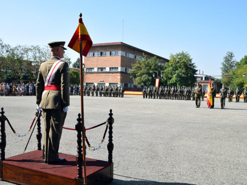Soldiers in uniform standing in formation with flags, a building in the background, and a clear blue sky.