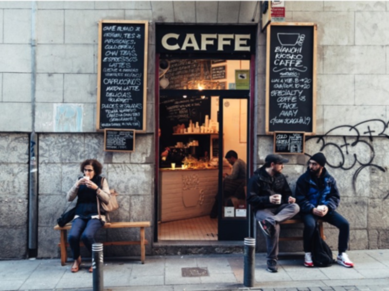 Personas disfrutando de café afuera de un café con menú de bebidas escritas en tablas de pizarra, en un entorno urbano.