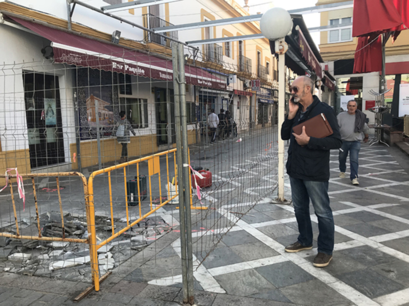 Hombre hablando por teléfono en una calle adoquinada con obras en construcción y negocios al fondo.