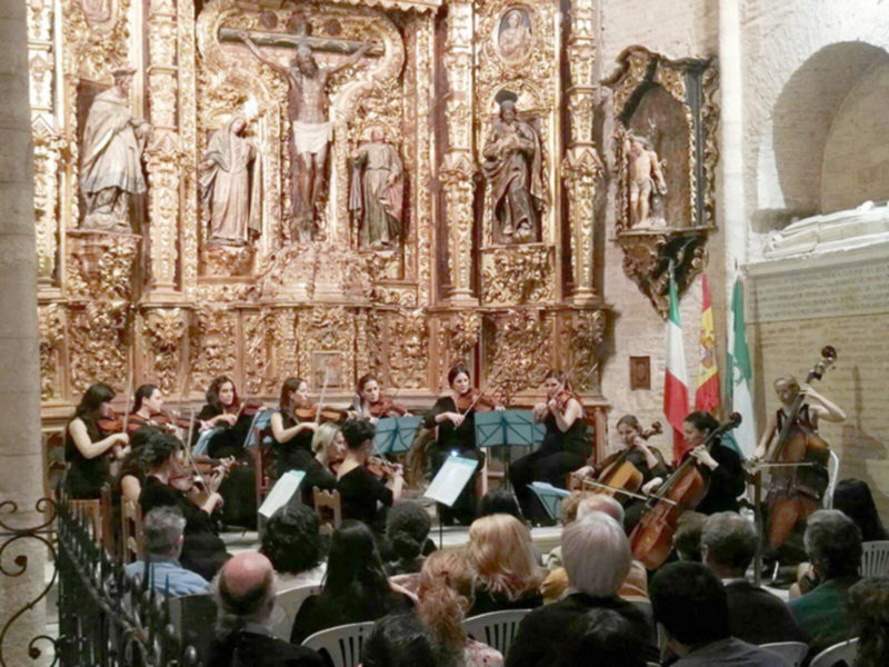 Orquesta de cámara en un concierto religioso en una iglesia con un altar dorado y estatuas religiosas.