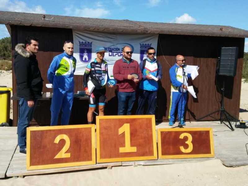 Ganadores de una carrera de ciclismo en una playa, con una bandera de la localidad en el fondo.