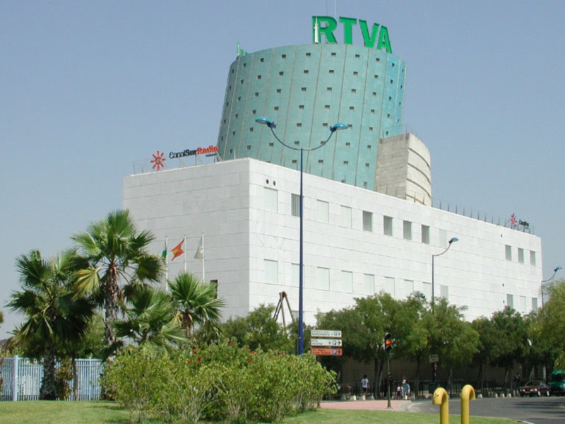 Edificio RTVA con torre circular y banderas en la parte superior, rodeado de árboles y jardines en un día soleado.