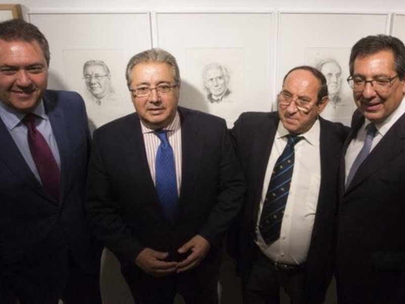 Tres hombres en trajes posan frente a una pared con retratos de personas. Todos llevan corbatas y parecen estar en una reunión formal.