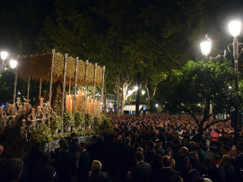 Procesión nocturna con una imagen procesional en un entorno urbano iluminado.
