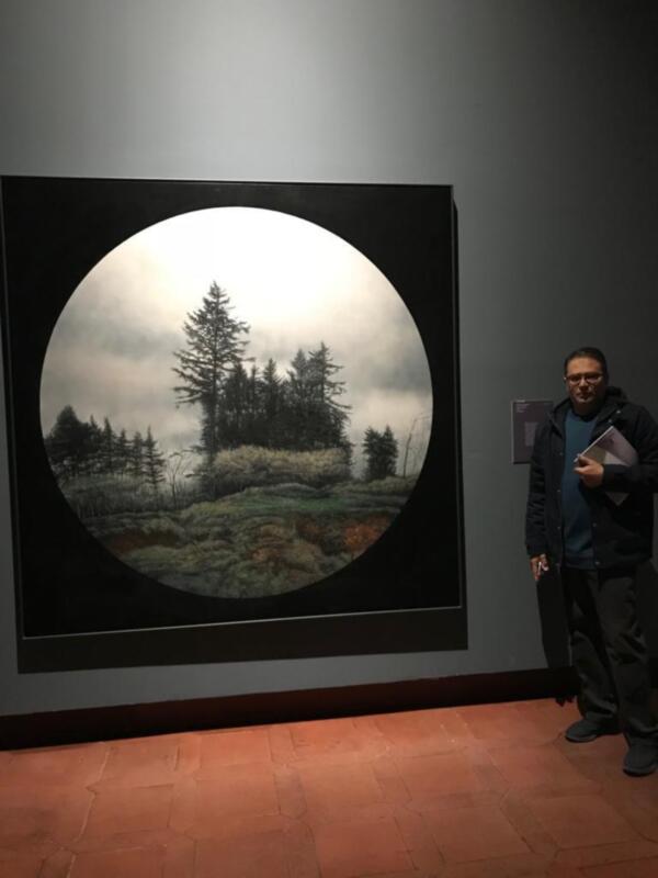 Artista junto a obra enmarcada con paisaje circular de bosque nebuloso.