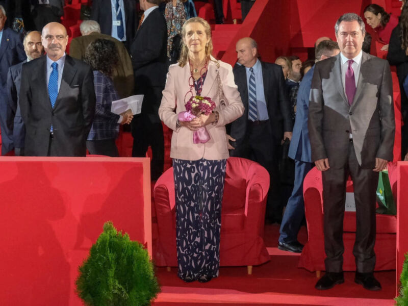 Persona destacada en un evento formal, rodeada de asistentes en un escenario con sofás rojos y plantas verdes.