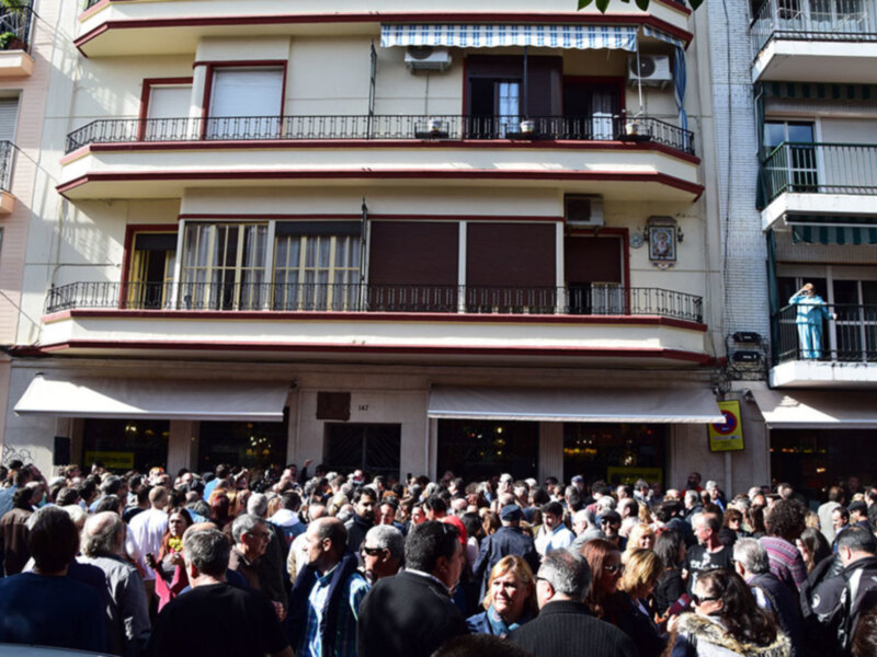 Gran multitud congregada frente a un edificio con balcones y tiendas en una calle urbana.