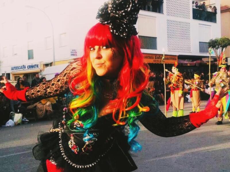 Una bailarina en un carnaval, con un vestido negro y una peluca roja, baila en la calle. El público observa desde la acera.