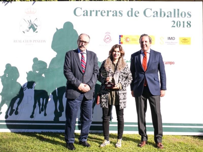 Premiación en la Carreras de Caballos 2018, con una mujer recibiendo un trofeo junto a dos hombres en trajes, frente a un fondo con el logo del evento y la bandera española.