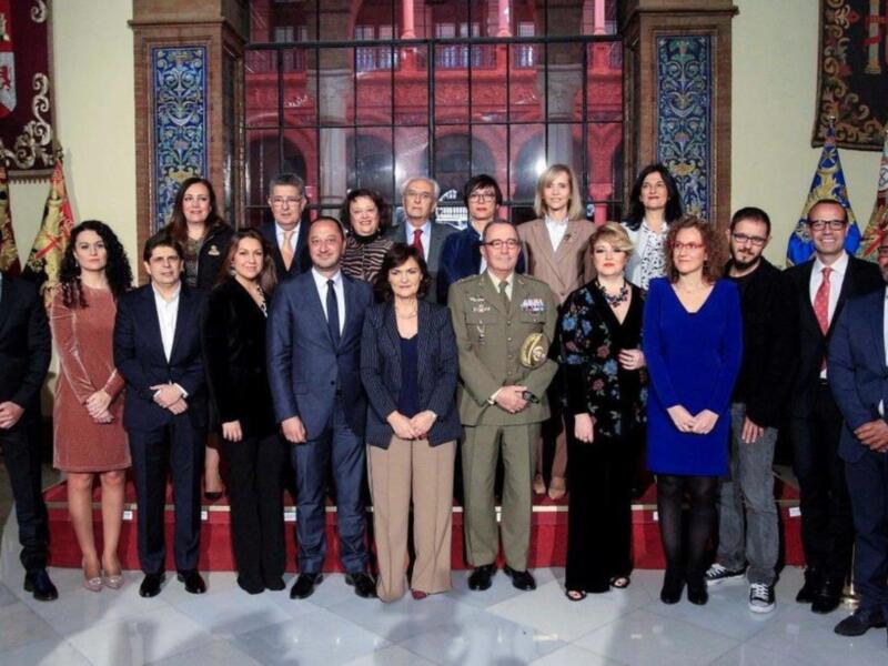 Grupo de personas posando en una sala con decoración clásica y banderas de España en el fondo.