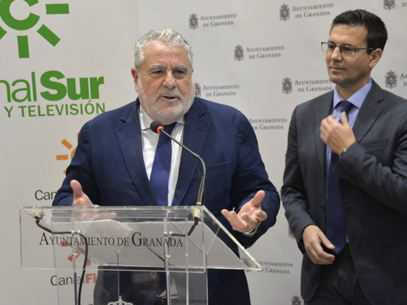 Dos hombres en trajes hablan en un evento de la radio y televisión de Granada. El fondo muestra el logotipo de Canal Sur y el Ayuntamiento de Granada.