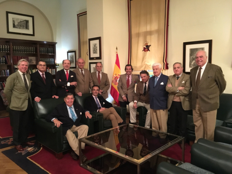 Grupo de hombres en trajes posando para una foto en un salón elegante con una bandera española en el fondo.