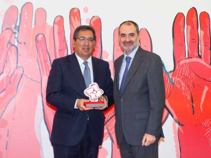 Dos hombres en trajes posan junto a un premio con una ilustración de manos en el fondo.