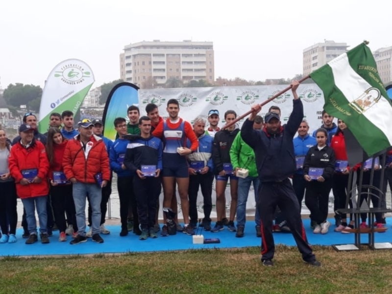 Equipo de remo celebrando en una ceremonia de premiación junto al río, con bandera y trofeos.