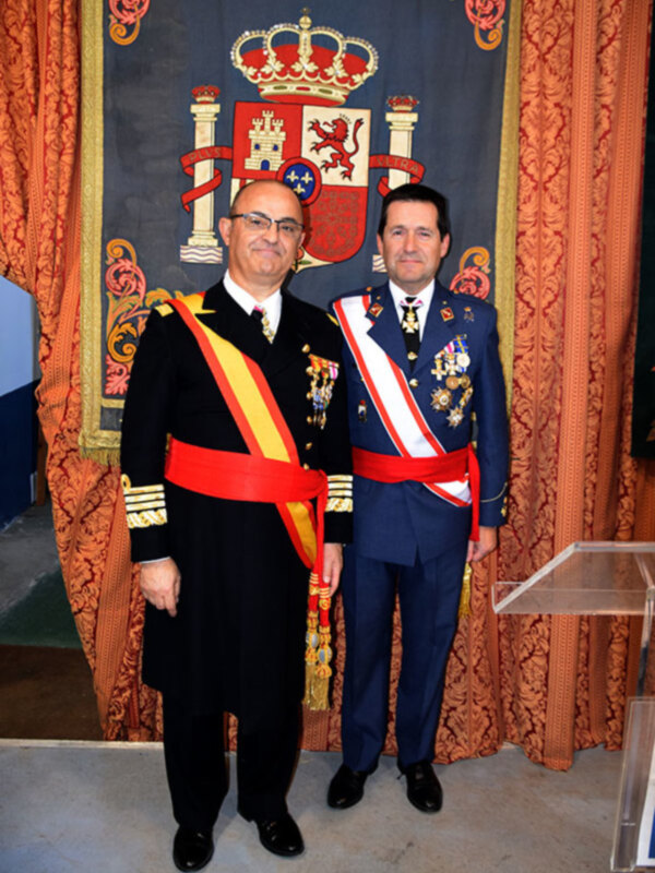 Dos hombres en uniforme militar posan frente a un escudo heráldico real, con un fondo de cortina decorada.