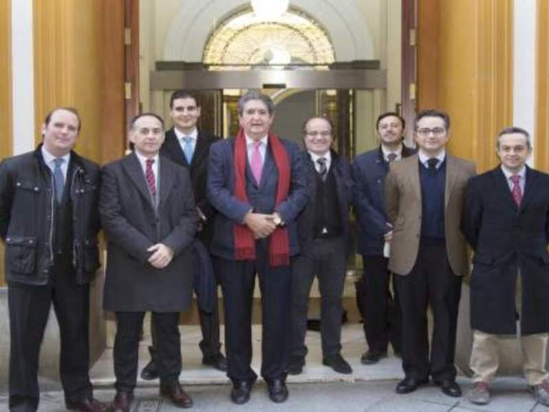 Grupo de hombres profesionales posando en la entrada de un edificio oficial, vestidos con trajes y bufandas, posiblemente en una reunión formal.