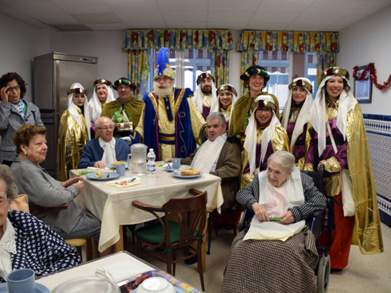 Un grupo de personas vestidas de trajes tradicionales se reúne en una sala decorada con adornos navideños, posiblemente para una celebración festiva. La mesa está servida con platos y vasos, y la atmósfera es festiva y amistosa.
