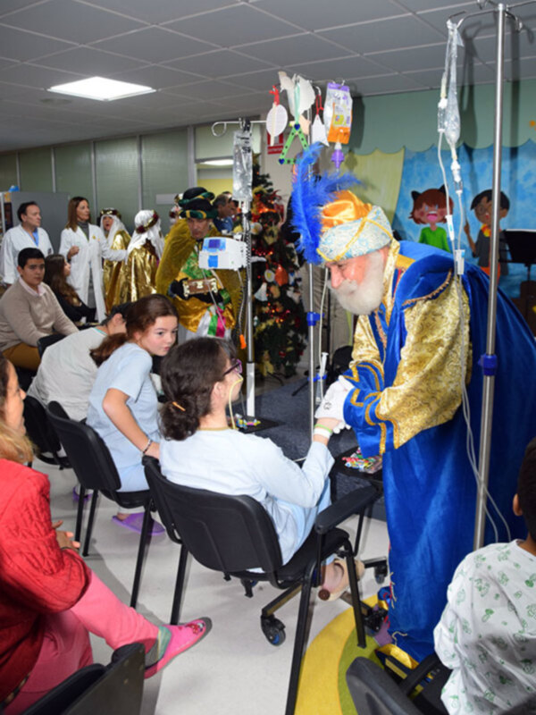 Un clásico mago en traje de circo interacciona con pacientes en una sala de hospital, rodeado de otros personajes y decoración festiva.