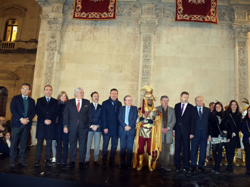 Gruppo di persone in abiti formali posano per una foto in un ambiente storico con colonne e arazzi rossi.