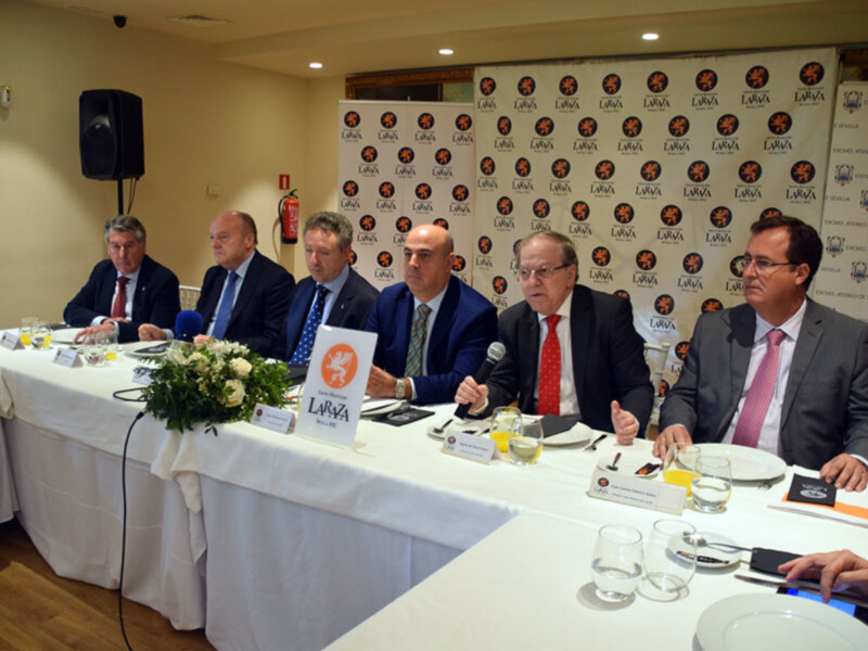 Grupo de personas sentadas en una mesa con bandejas, copas y un microfono, en una conferencia o reunión formal.