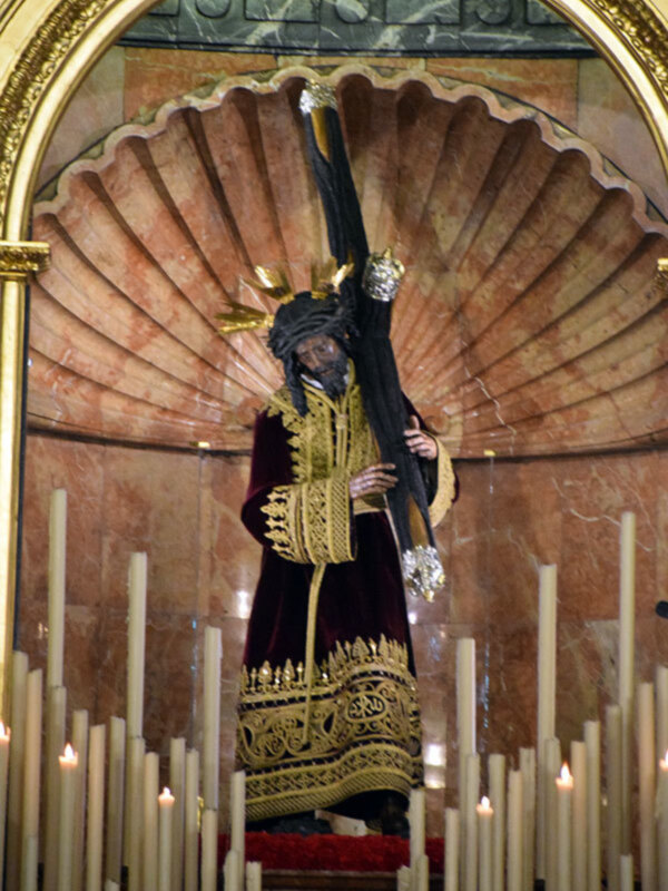Figura de Jesús Nazareno en un altar con velas encendidas, vestido con traje rojo y dorado, en un entorno de mármol rojo.