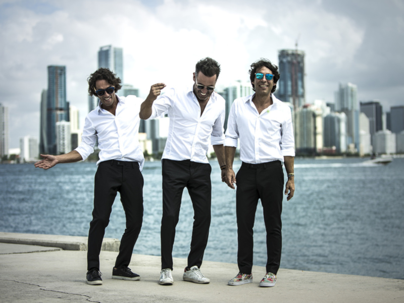 Tres hombres en camisas blancas y pantalones negros posan juntos en un muelle frente al horizonte urbano de Miami.