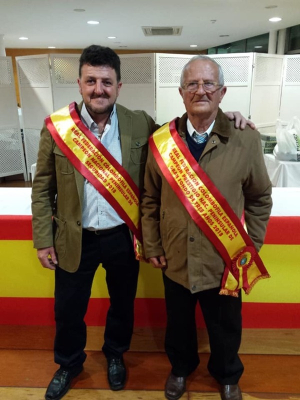 Dos personas posando juntas en un evento, uno con un cinturón amarillo y rojo con texto en español, posiblemente en un evento deportivo o de reconocimiento.