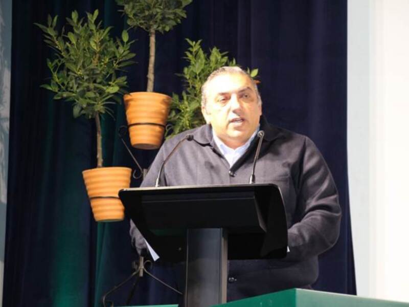 Persona hablando desde un podio en un evento formal, con plantas en macetas en el fondo.
